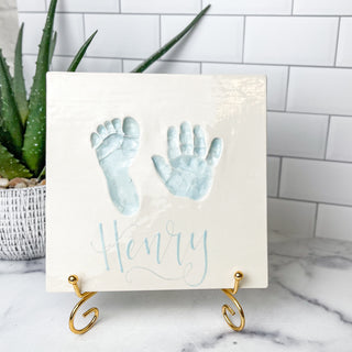 White square plate with blue impressions of a child's hand and footprint. 